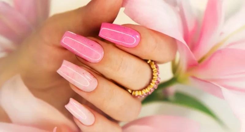 Pink acrylic nails with a glossy finish held delicately against a pink lily flower, showcasing a feminine and floral-inspired manicure with a touch of sparkle.