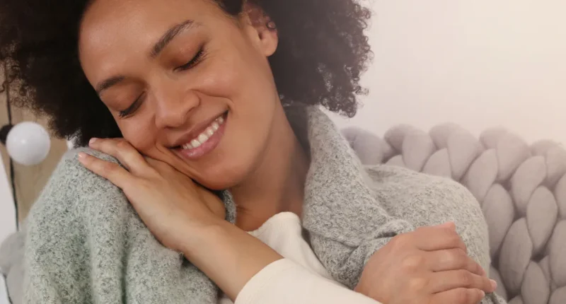 A smiling woman with curly hair hugging herself on a cozy couch, depicting self-love and comfort.