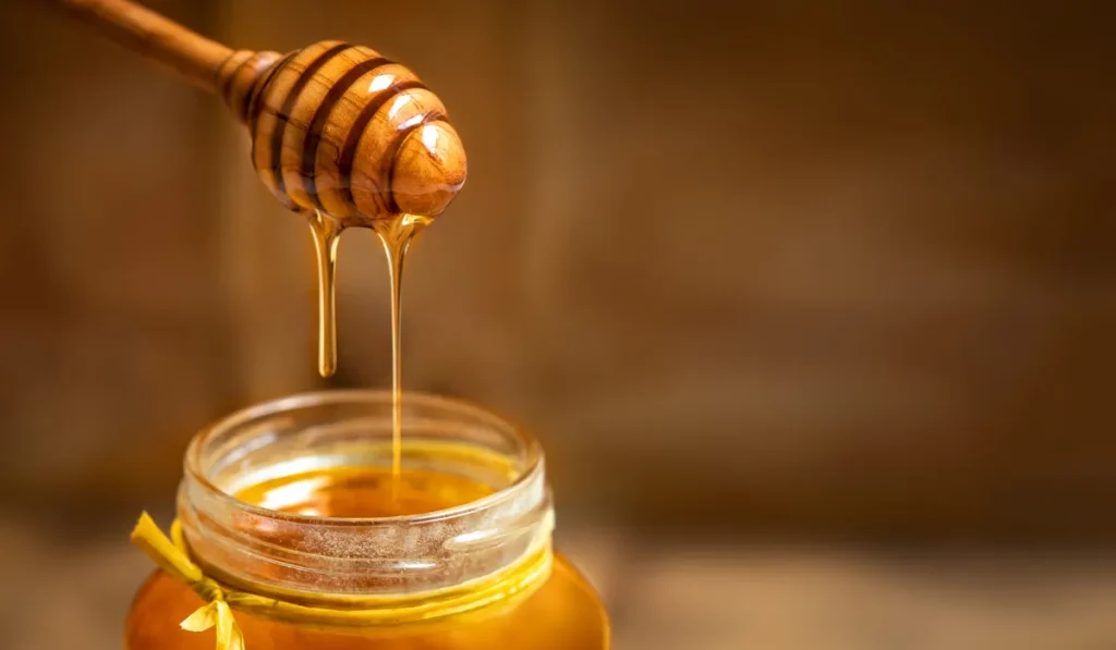06 Natural Ways to Get Rid of Pimples 2024 6 A close-up of a wooden honey dipper dripping golden honey into a jar, representing natural ingredients for skincare or healthy eating.