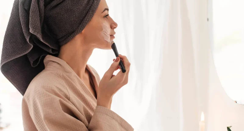 Serene woman in a soft robe applying foundation, standing by a window with sheer curtains, showcasing a part of a daily beauty ritual with natural light