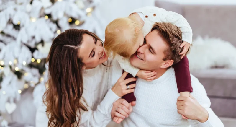 A joyful family with a toddler celebrating Christmas indoors, with the mother kissing the child's cheek and the father smiling broadly in a cozy, festive room with a white Christmas tree illuminated by soft lights.