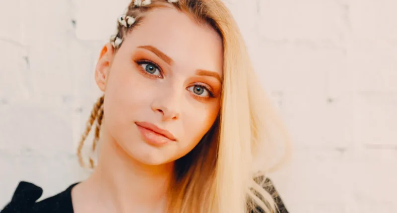 Portrait of a young woman with a side braid adorned with white flowers, her blonde hair contrasted against a light, textured background, highlighting a fresh and youthful look