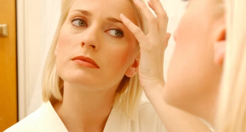 A concerned middle-aged woman with blonde hair examines her facial skin for signs of aging or wrinkles, standing in a well-lit bathroom with a neutral expression