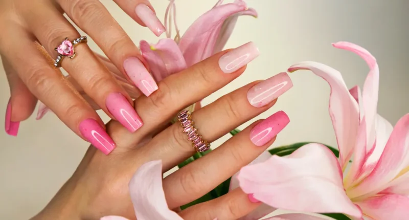 A harmonious blend of pink ombre coffin nails complemented by delicate lily petals, showcasing various shades of pink that transition smoothly from a deep rose at the tips to a soft blush near the cuticles. The hands are adorned with elegant jewelry, including a ring with a pink heart-shaped gemstone and a band of pink gems, enhancing the feminine charm of the nail art.