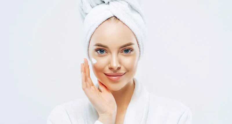 Woman with a towel on her head applying a dab of cream to her cheek, embodying a refreshing spring skincare routine with a focus on moisturization