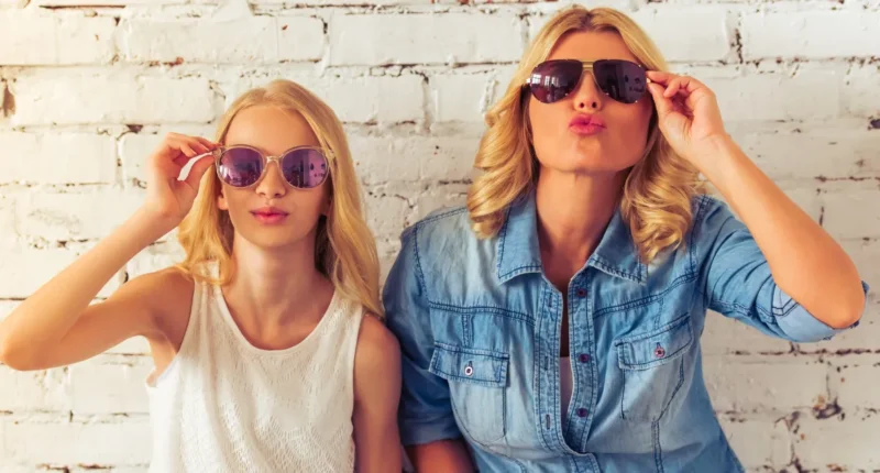 A fashionable mother and daughter duo playfully wearing oversized sunglasses against a white brick wall, embodying a fun and trendy family look for a featured style blog image 2024.