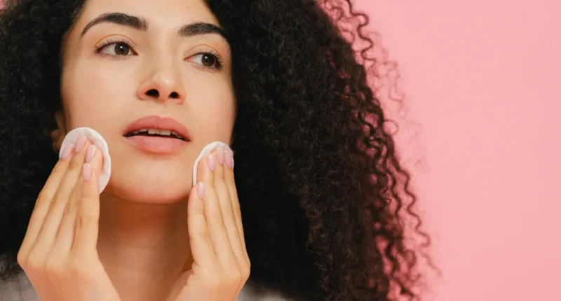 Curly-haired woman with a thoughtful expression gently cleanses her face with cotton pads against a soft pink background, highlighting a daily skincare routine.