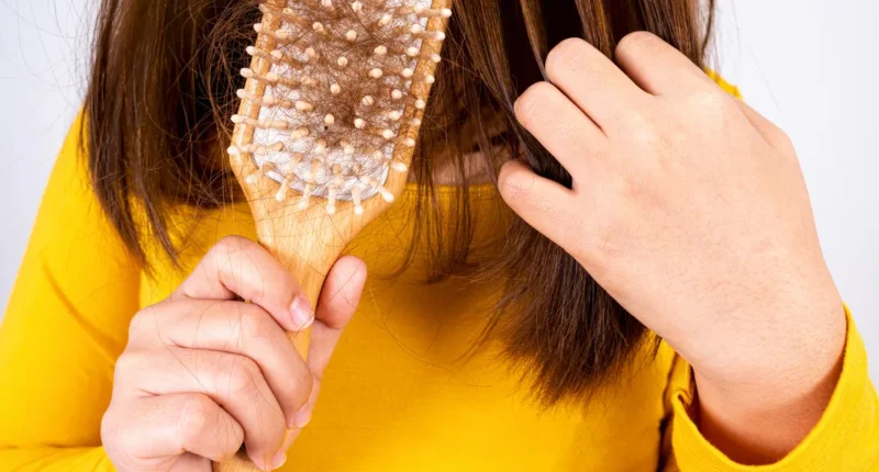 A person in a white top holding a hairbrush with strands of hair, indicating hair fall, with a concerned expression on their face