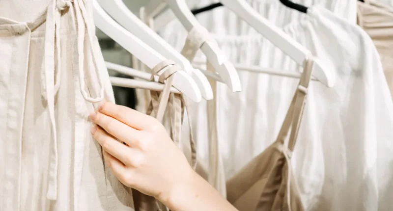 Close-up of a hand selecting garments on wooden hangers, focusing on sustainable fashion choices in a light, clean retail space, reflecting 2024's green wardrobe revolution