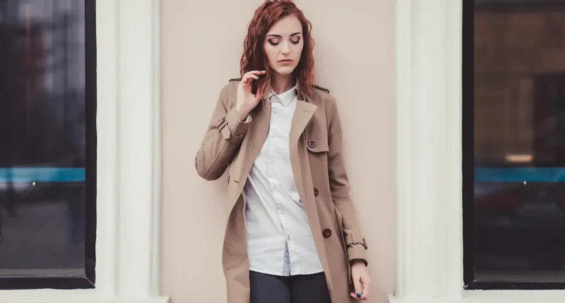 Fashionable woman in a classic beige trench coat with a white shirt, deep in thought, standing against a cream-colored wall with a city backdrop.