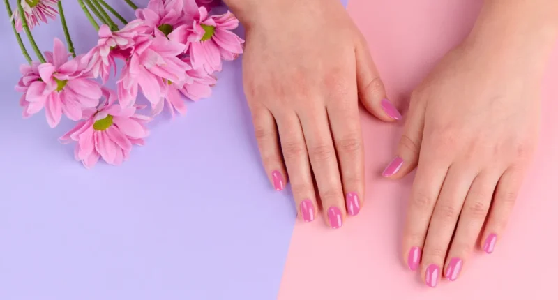 Two hands with nails polished in a radiant pink hue, resting on a dual-tone background of lilac and pink, complemented by soft pink spring flowers