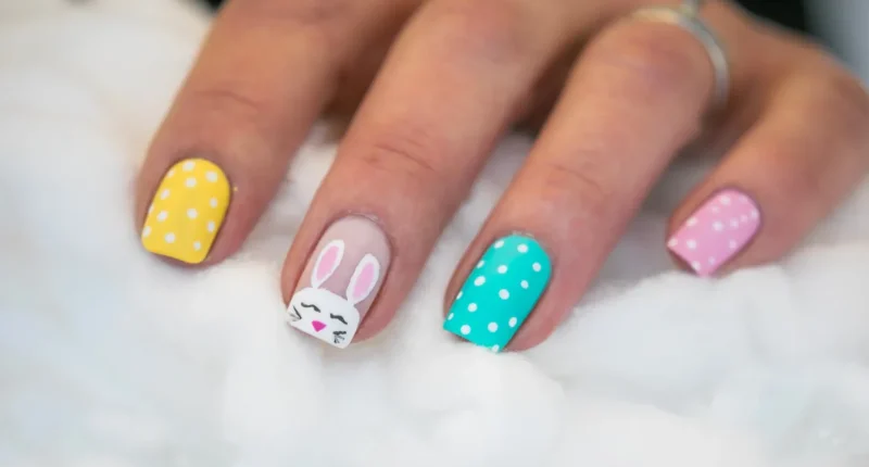 A hand rests on a plush white surface showcasing a colorful Easter nail design with bright yellow and turquoise polka-dotted nails, one featuring a cute white bunny face with pink ears for a festive spring look.