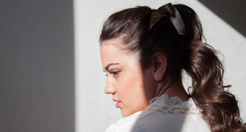 Woman captured in a contemplative moment with her hair styled in an elegant ponytail secured with a bow accessory, radiating a vintage charm in a modern setting.